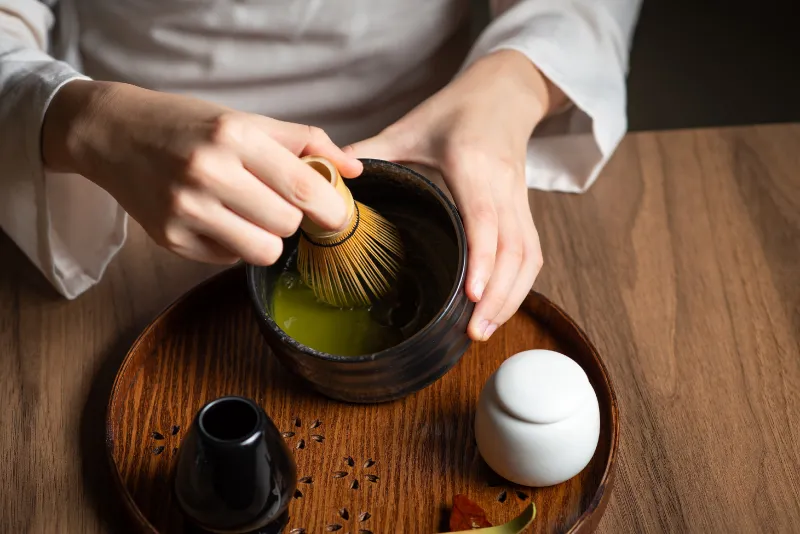 Hands whisking matcha with a bamboo chasen in a dark tea bowl on a round wooden tray